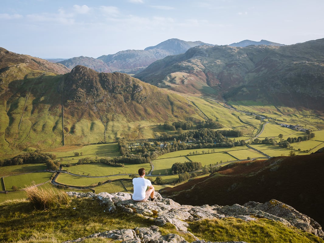 Man sits on the top of a mountain overlooking a green valley