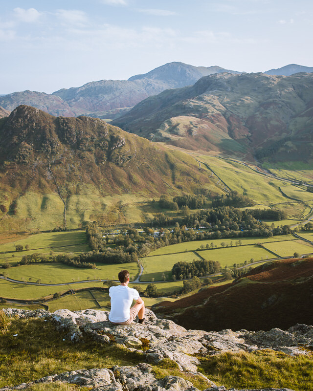 Man sits on the top of a mountain overlooking a green valley