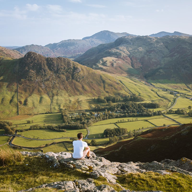 Man sits on the top of a mountain overlooking a green valley