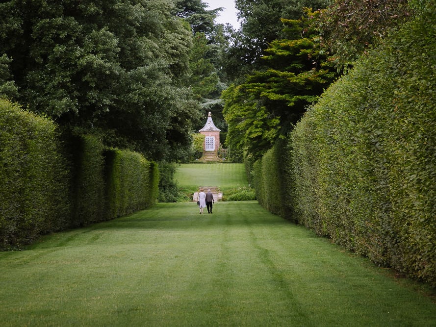 Couple walk over manicured lawn between high hedges