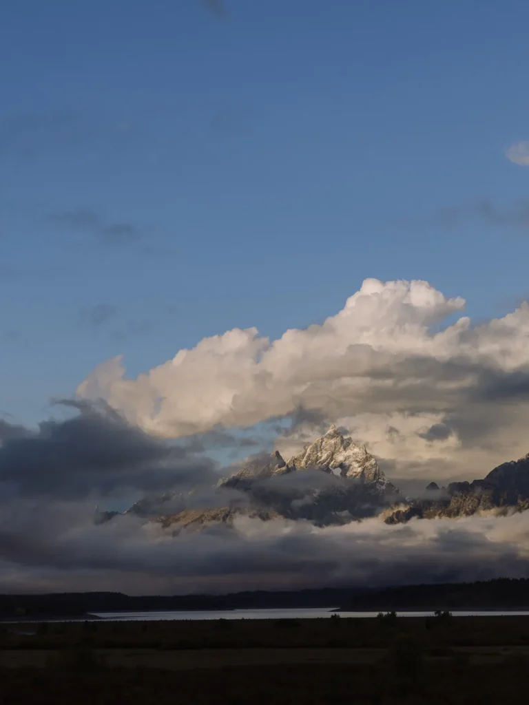 Grand Teton peaks partly obscured by clouds and mist above a dark valley at sunrise.