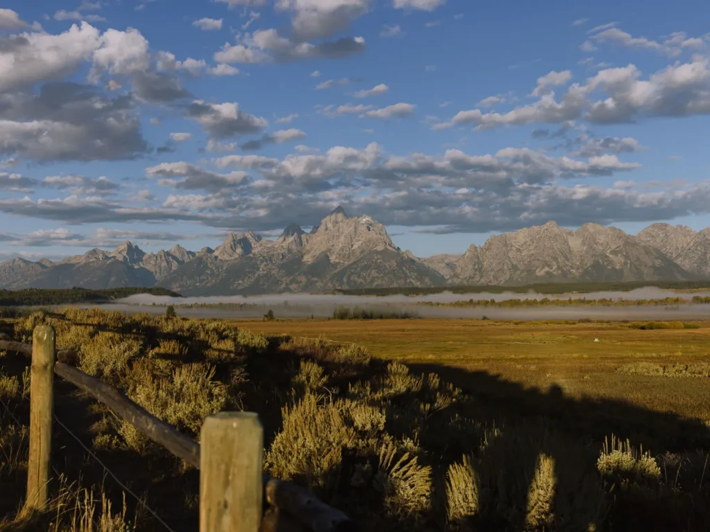 Grand Teton mountains lit by warm sunrise light above a grassy plain under scattered clouds.