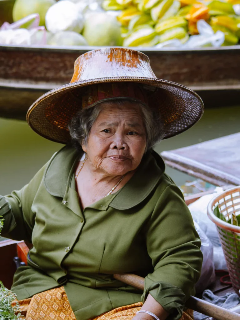 An elderly female fruit vendor wearing a traditional Thai bamboo hat sits in her boat at Damnoen Saduak floating market.