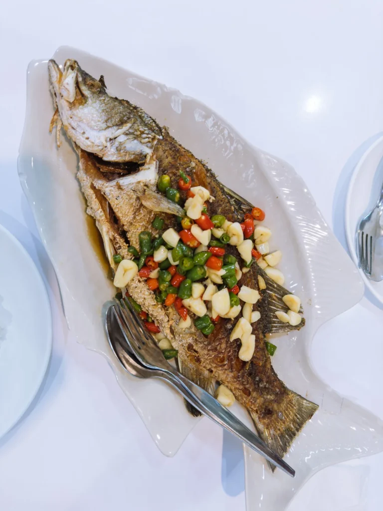 A whole deep-fried fish topped with chopped garlic, red and green chillies, served on a white platter at a Bangkok restaurant.