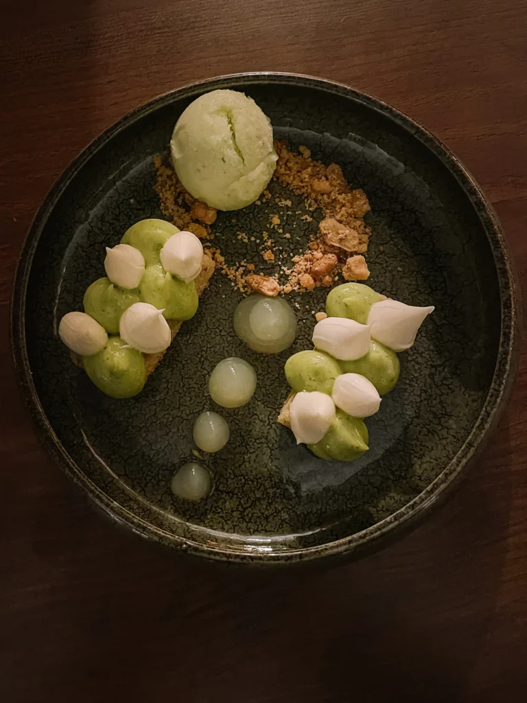 An elegantly plated green dessert with a scoop of matcha ice cream, piped cream, translucent spheres, and crumble on a dark ceramic bowl.