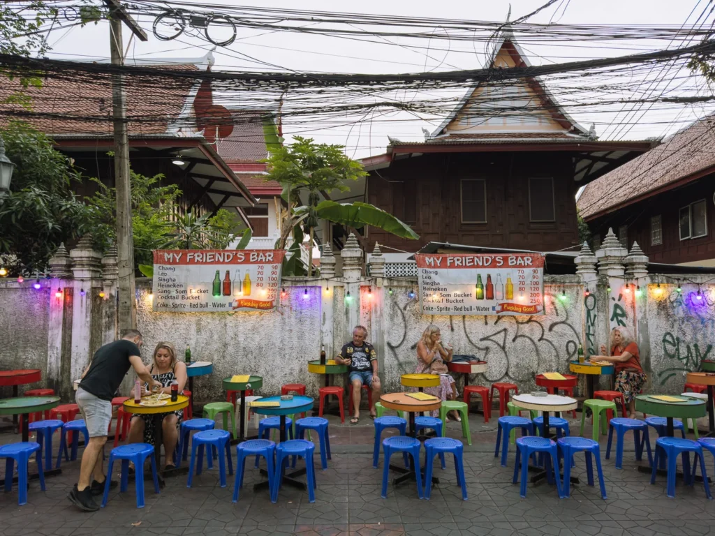 Tourists sit at colourful plastic tables and stools outside My Friend's Bar, a casual open-air bar on Khao San Road, Bangkok.
