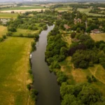 Aerial view of the River Thames winding through the Oxfordshire countryside at Clifton Hampden, with a narrowboat on the water, golden meadows on one side and the village nestled among lush green trees on the other