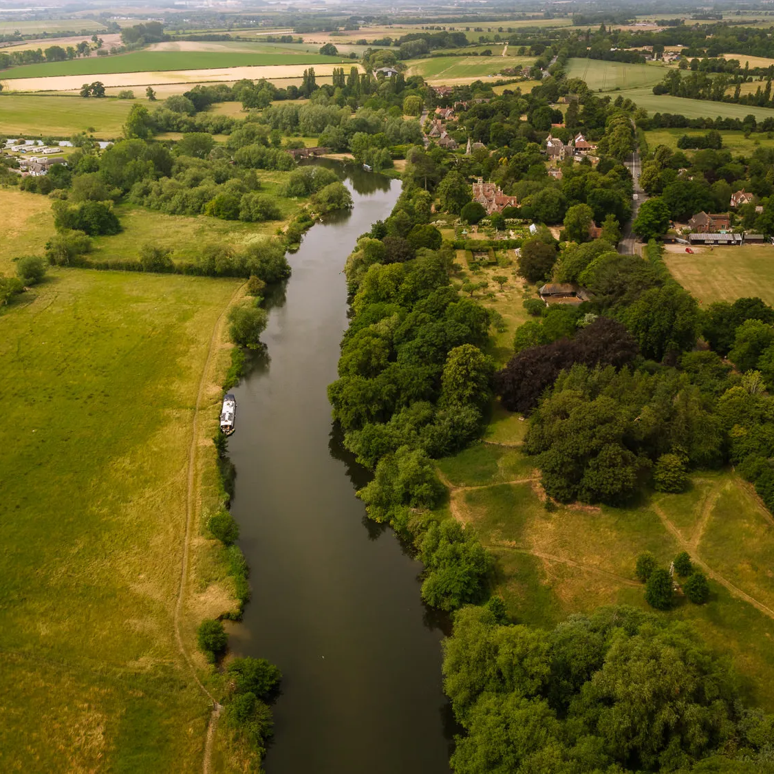 Aerial view of the River Thames winding through the Oxfordshire countryside at Clifton Hampden, with a narrowboat on the water, golden meadows on one side and the village nestled among lush green trees on the other
