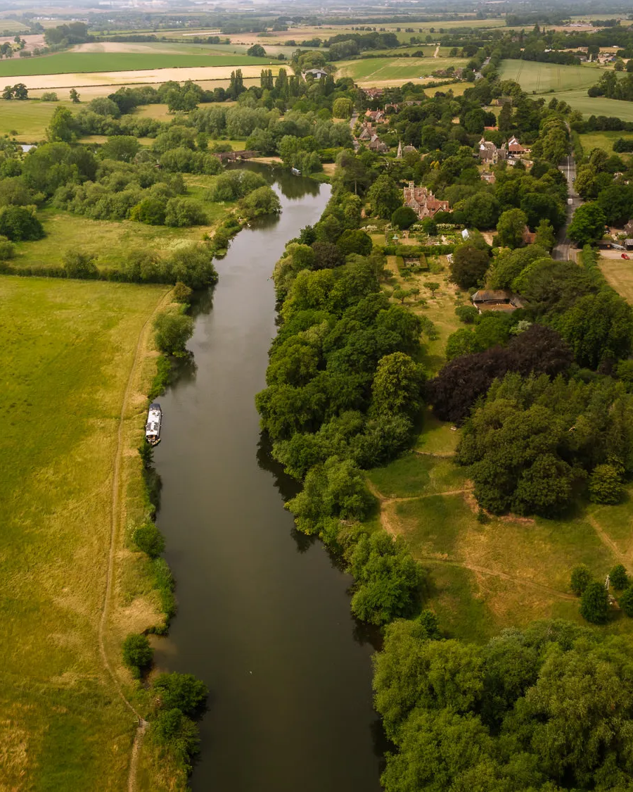 Aerial view of the River Thames winding through the Oxfordshire countryside at Clifton Hampden, with a narrowboat on the water, golden meadows on one side and the village nestled among lush green trees on the other