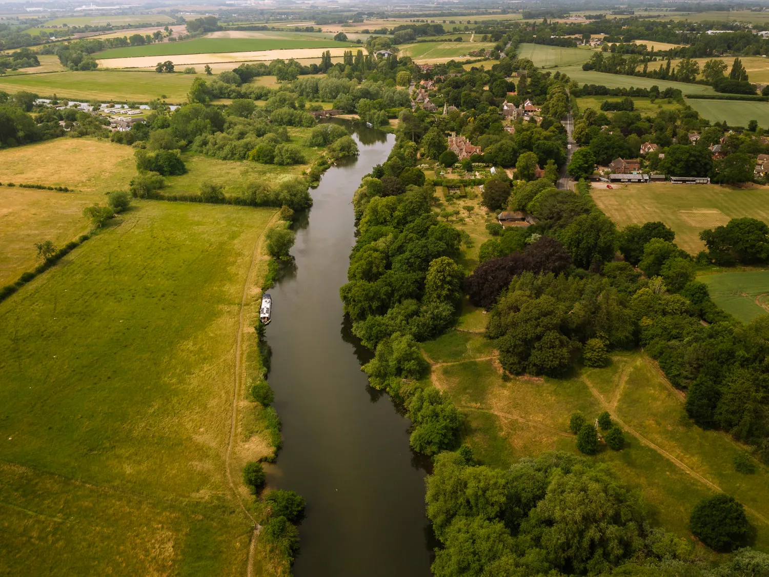 Aerial view of the River Thames winding through the Oxfordshire countryside at Clifton Hampden, with a narrowboat on the water, golden meadows on one side and the village nestled among lush green trees on the other