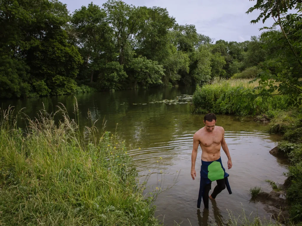 A wild swimmer wading out of the River Thames at Clifton Hampden, holding a swim cap, with lily pads and lush green riverbank vegetation in the background