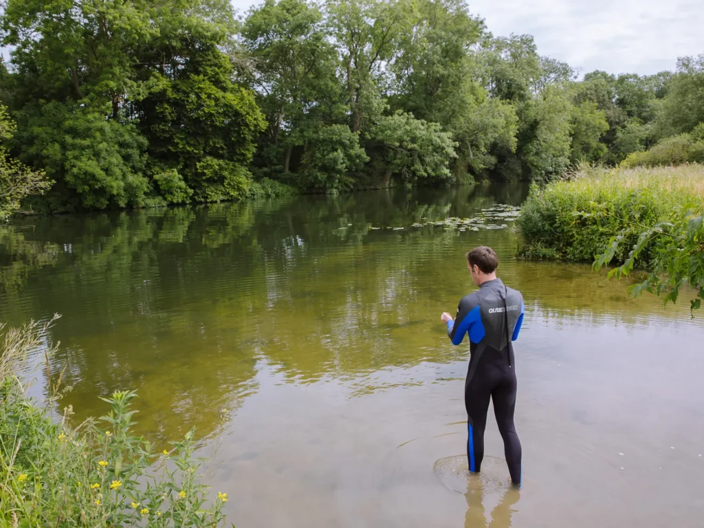 A wild swimmer in a wetsuit wading into the River Thames at Clifton Hampden, looking out across the calm green water with lily pads and lush woodland lining the far bank