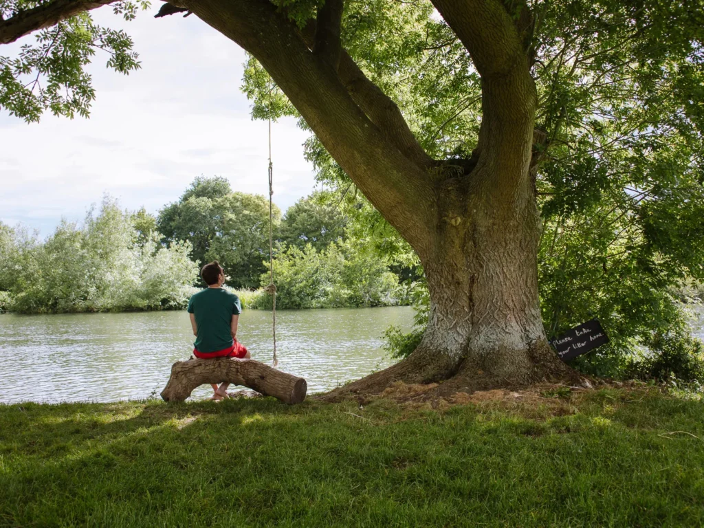 A person sitting on a fallen log beneath a large tree beside the River Thames at Moulsford, with a rope swing hanging overhead and a small handwritten "please take your litter home" sign propped against the trunk
