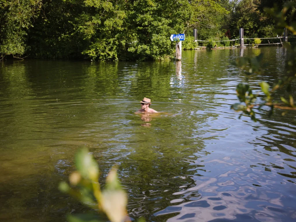 A wild swimmer floating in the River Thames at Shiplake wearing a straw hat, surrounded by lush green trees and a navigation sign on the river bank behind
