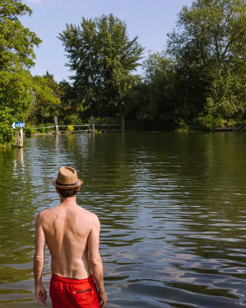 A wild swimmer wading into the River Thames at Shiplake wearing red swim shorts and a straw hat, looking out across the calm green water towards the tree-lined far bank