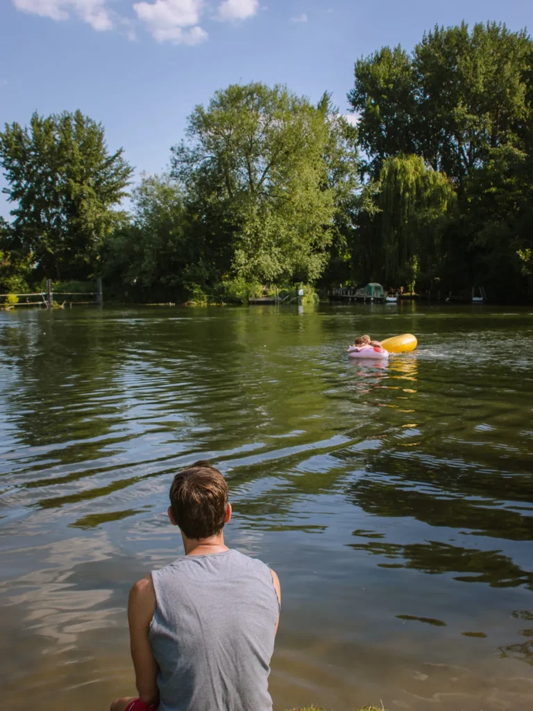 A person sitting at the water's edge watching a child float on an inflatable in the River Thames at Shiplake on a sunny summer's day, with willow trees and a green riverbank in the background