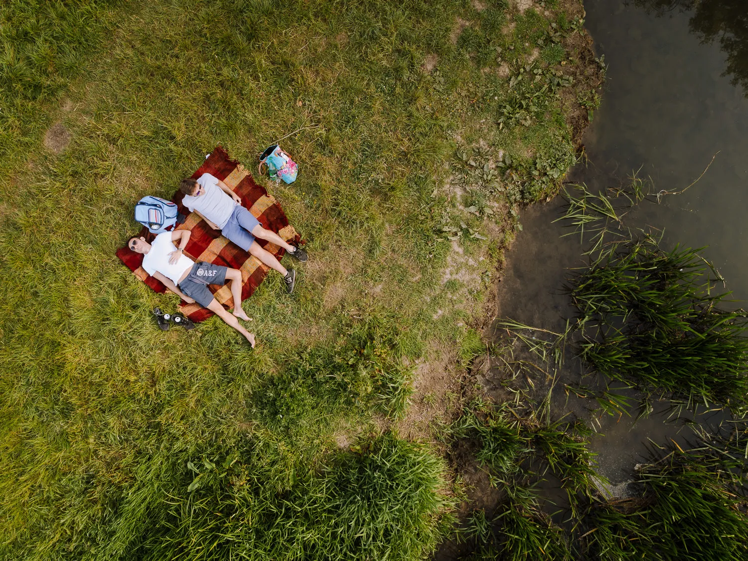 Aerial view of a couple lying on a picnic blanket on the grassy bank of the River Windrush at Minster Lovell, with reeds and the calm river visible beside them on a sunny summer's day