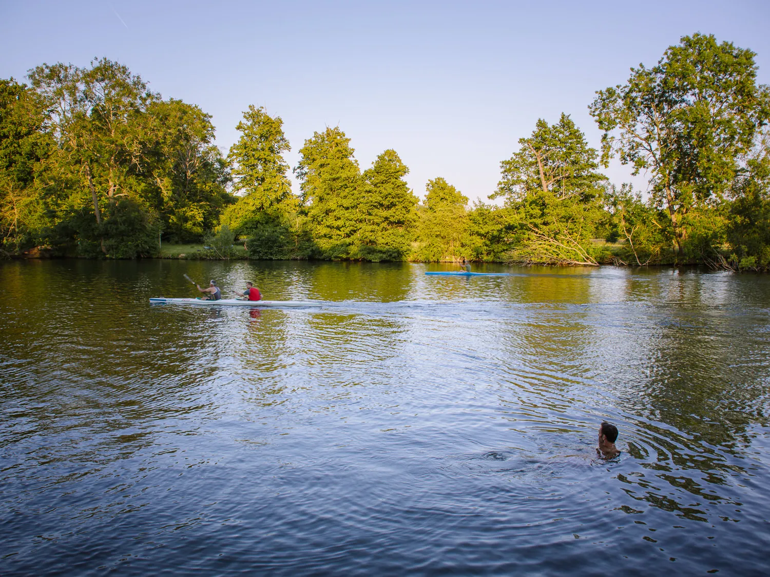 A wild swimmer in the River Thames at Medmenham alongside kayakers paddling past, with lush green trees lining the far bank in warm evening light