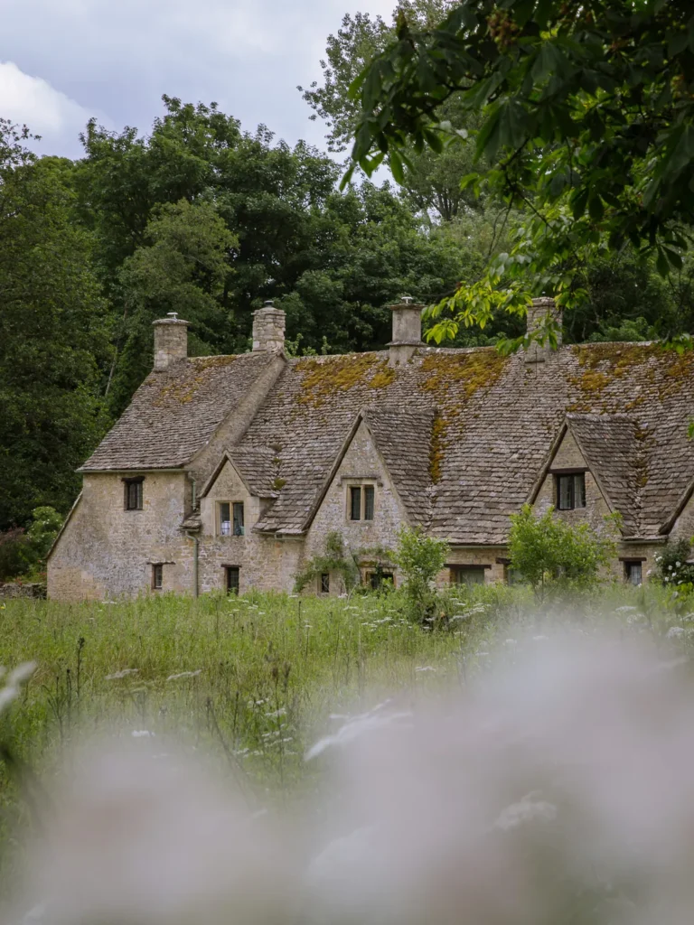 Historic stone cottages at Arlington Row in Bibury, surrounded by trees and wildflowers in the Cotswolds.