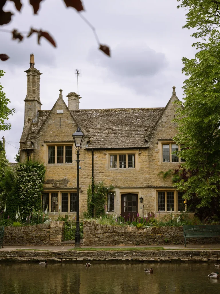 Honey-coloured stone house beside the River Windrush in Bourton-on-the-Water, with ducks on the water and a traditional street lamp in front.