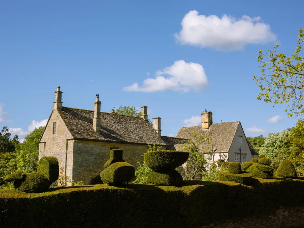 Cotswold stone cottages and sculpted topiary gardens in Chipping Campden under a bright blue sky.