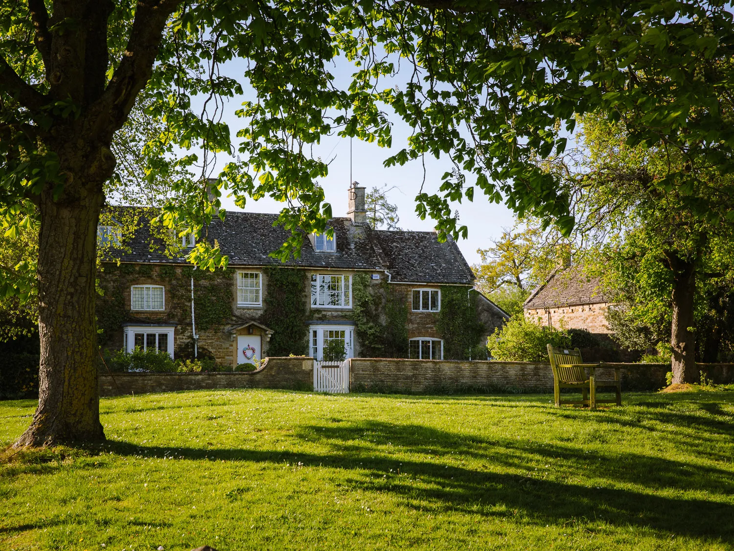 Traditional stone cottages overlooking a leafy village green with benches in Kingham, Cotswolds.
