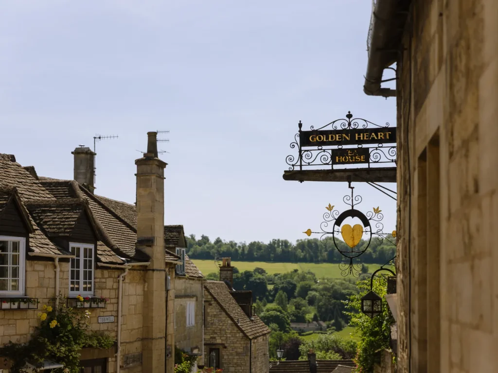Golden Heart pub sign above a Cotswold stone street in Painswick, with countryside views beyond.