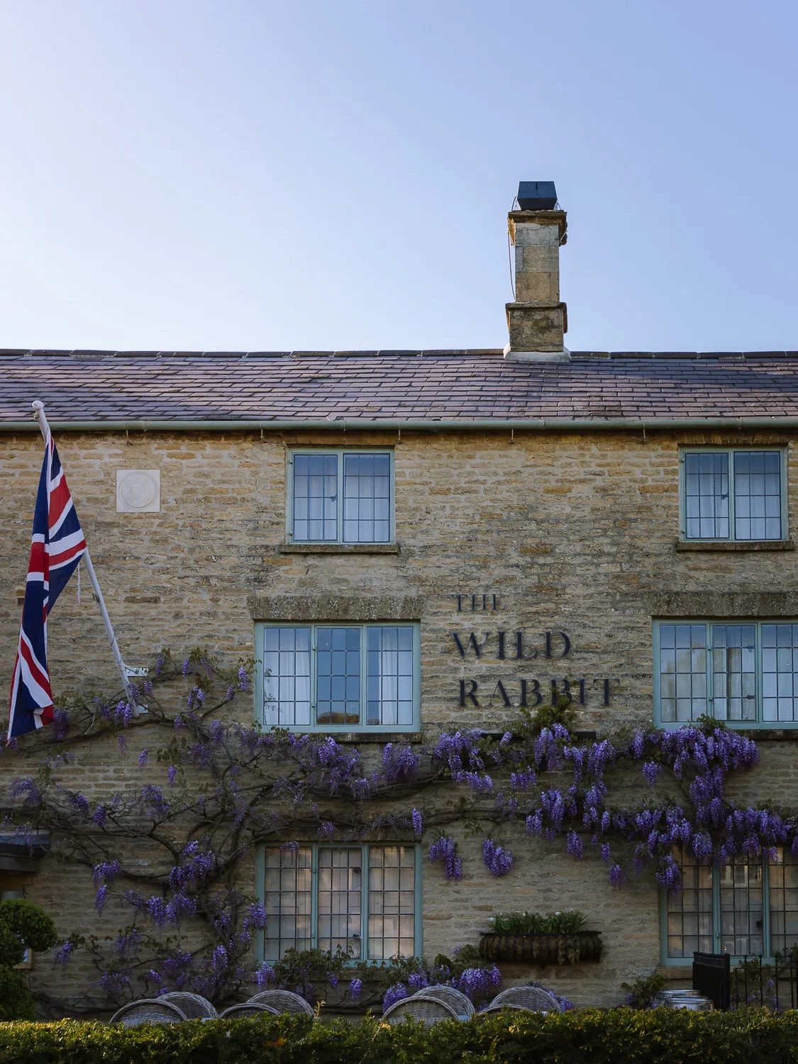 The Wild Rabbit in Kingham, a Cotswold stone pub covered in purple wisteria with a Union Jack outside.