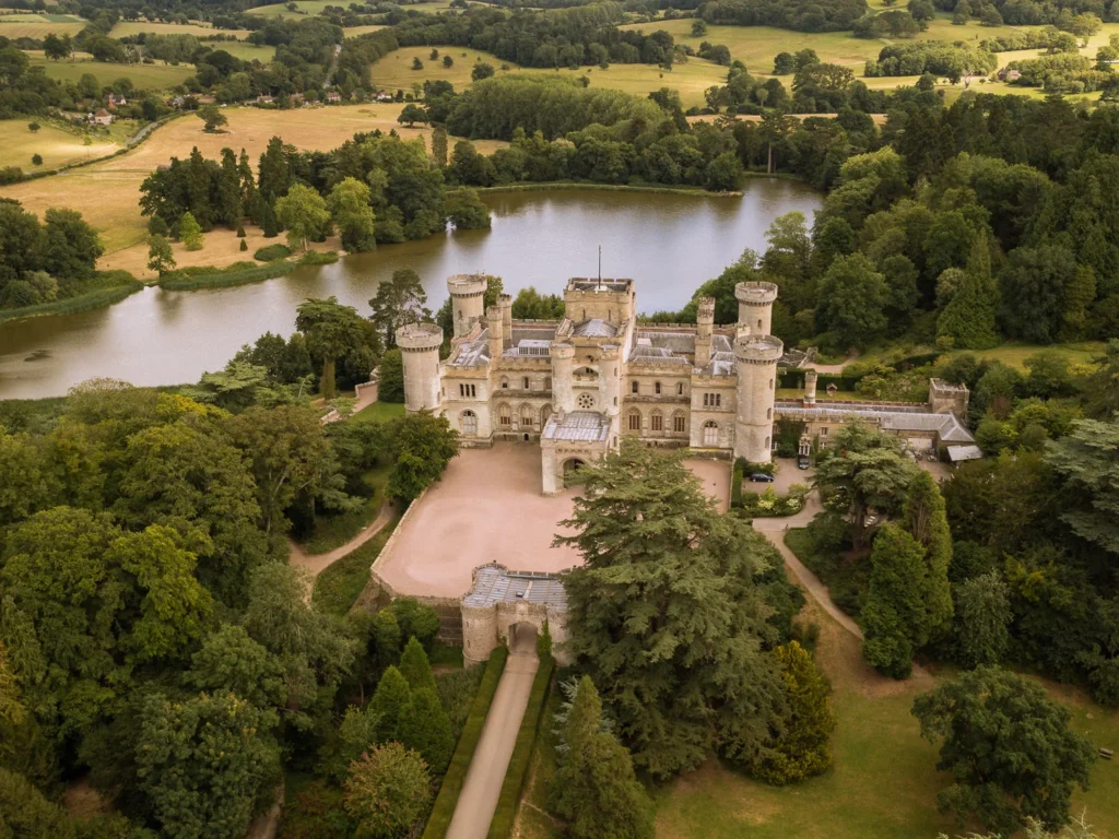 An aerial drone view of Eastnor Castle, a historic Gothic Revival stone fortress with round turrets and a central keep, surrounded by a large lake, dense green parkland, and a spacious gravel courtyard approached by a tree-lined driveway.