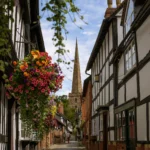 A sunny view looking down the cobbled Church Lane in Ledbury, framed by traditional black-and-white half-timbered buildings. A lush, brightly colored hanging flower basket dominates the left foreground, while the tall stone spire of St. Michael and All Angels Church rises at the end of the street under a blue sky with scattered clouds. Two people are walking in the distance.