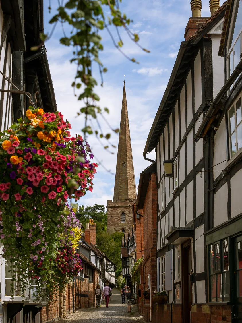 A sunny view looking down the cobbled Church Lane in Ledbury, framed by traditional black-and-white half-timbered buildings. A lush, brightly colored hanging flower basket dominates the left foreground, while the tall stone spire of St. Michael and All Angels Church rises at the end of the street under a blue sky with scattered clouds. Two people are walking in the distance.