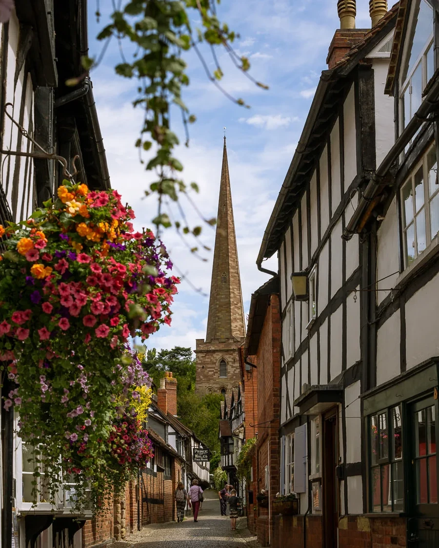 A sunny view looking down the cobbled Church Lane in Ledbury, framed by traditional black-and-white half-timbered buildings. A lush, brightly colored hanging flower basket dominates the left foreground, while the tall stone spire of St. Michael and All Angels Church rises at the end of the street under a blue sky with scattered clouds. Two people are walking in the distance.