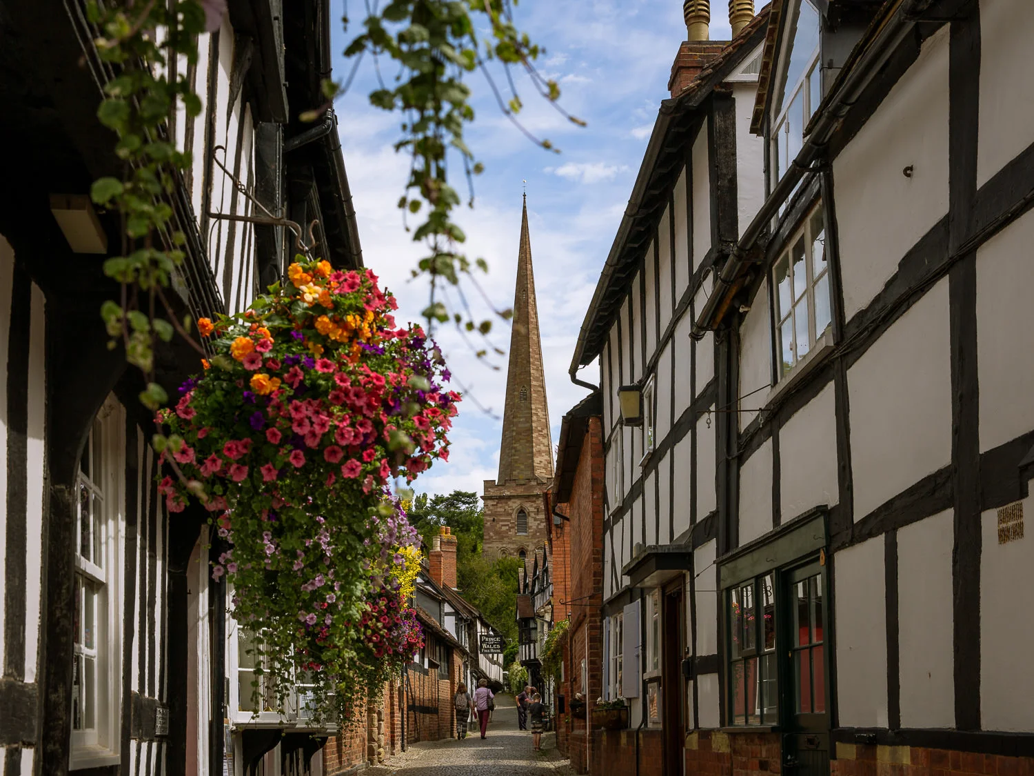 A sunny view looking down the cobbled Church Lane in Ledbury, framed by traditional black-and-white half-timbered buildings. A lush, brightly colored hanging flower basket dominates the left foreground, while the tall stone spire of St. Michael and All Angels Church rises at the end of the street under a blue sky with scattered clouds. Two people are walking in the distance.