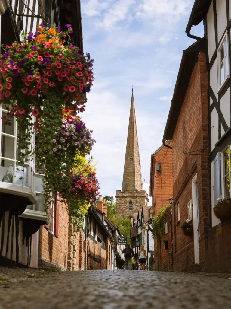 A low-angle, ground-level perspective looking up the narrow, cobbled Church Lane in Ledbury, flanked by black-and-white timber-framed buildings decorated with colorful hanging flower baskets. The tall stone spire of St. Michael and All Angels Church rises in the background