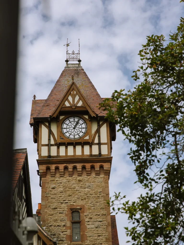 A low-angle close-up of the Victorian-era Ledbury Clock Tower, featuring a sandstone stone base and a white half-timbered upper section with a circular clock face, topped with a red-tiled roof and a decorative metal finial, partially framed by leaves.