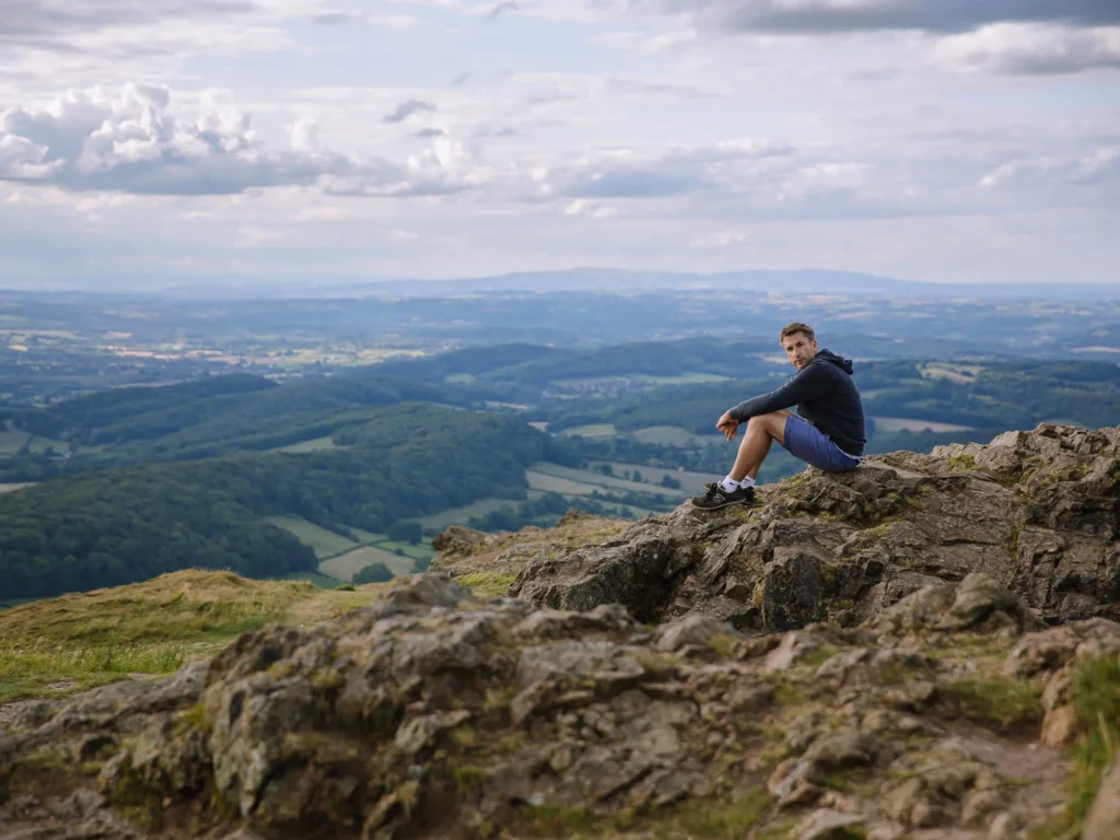A young man sitting on a rugged, rocky cliff edge, looking back at the camera while enjoying a panoramic view over rolling green hills and distant valleys of the Malvern Hills area.