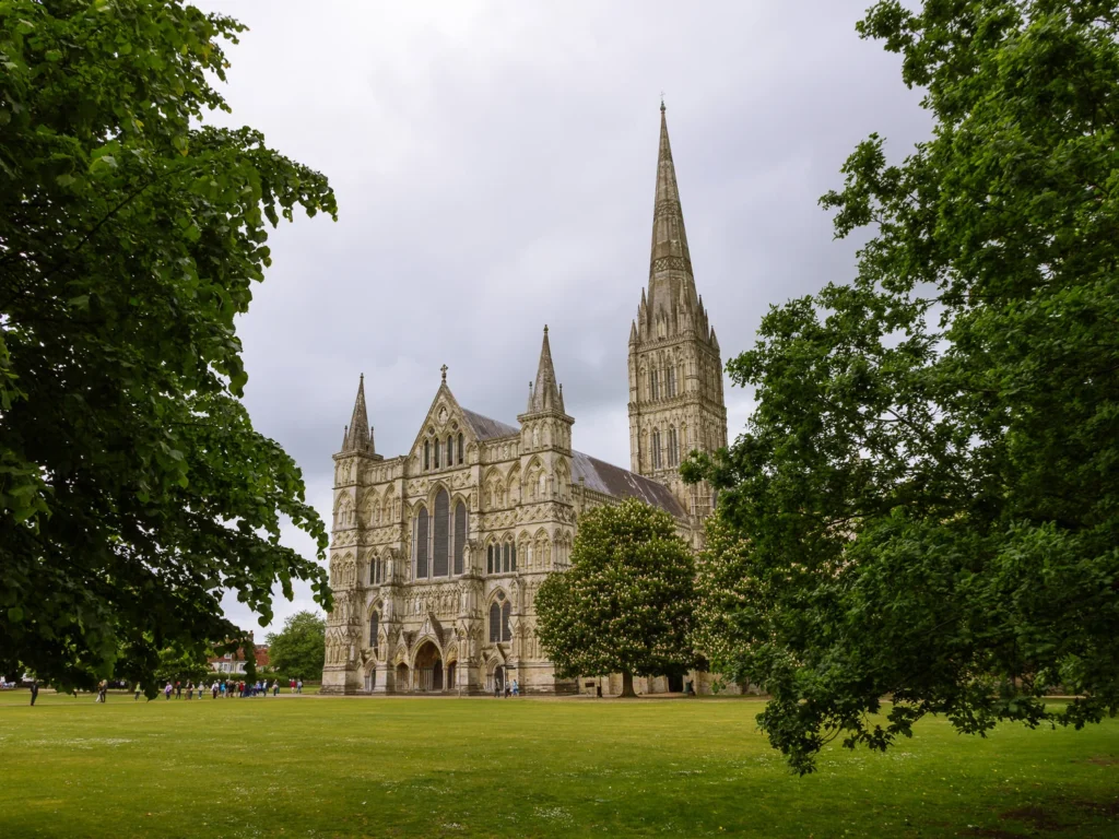 Salisbury Cathedral seen across a green lawn and framed by trees, with its tall spire rising above the cathedral building.