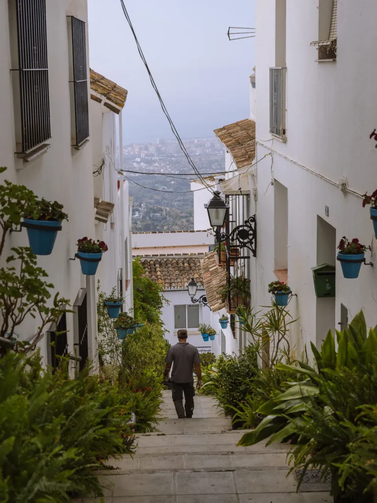 A narrow staircase street in Mijas Pueblo lined with blue flower pots and lush plants, with views of the Costa del Sol coastline stretching out below