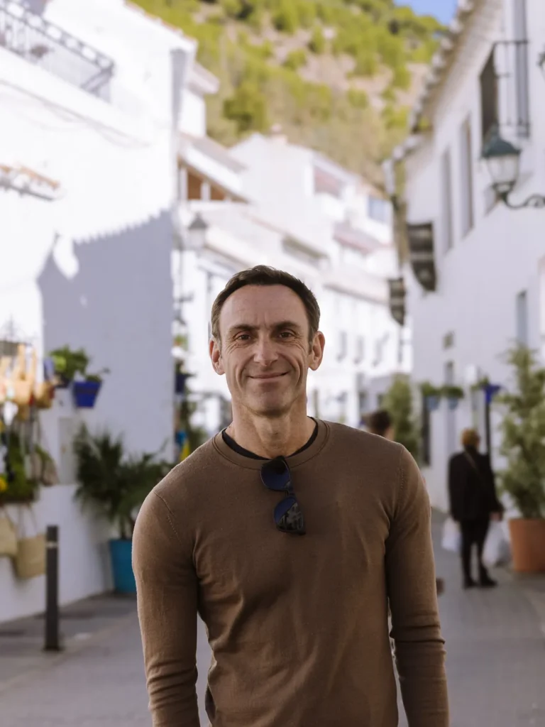 Mark standing on a whitewashed street in Mijas Pueblo, with blue flower pots, green hillside and traditional Andalusian buildings in the background
