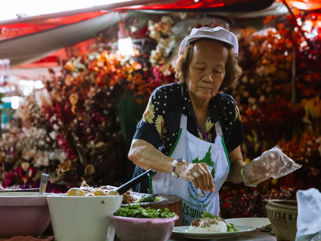 Elderly vendor seasons food amid vibrant flowers at Chatuchak Weekend Market in Bangkok.