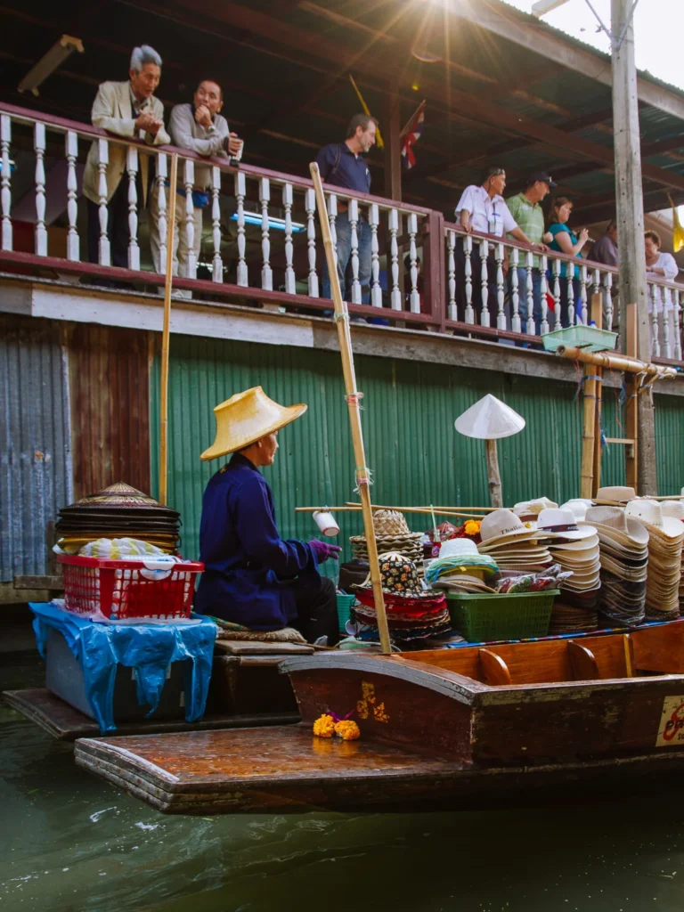 Vendor in conical hat sells hats and goods from boat at Damnoen Saduak Floating Market, with onlookers on riverside balcony.