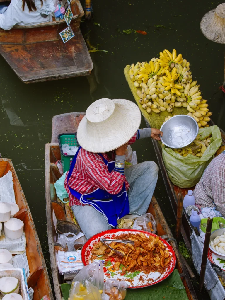 Floating market vendor in conical hat sells bananas and fried snacks from longtail boat at Damnoen Saduak