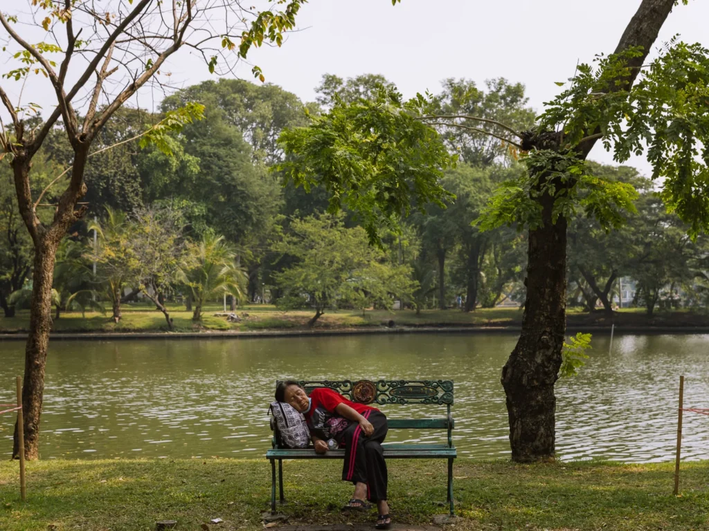 A person napping on an ornate green park bench beside a calm lake surrounded by lush trees in Lumpini Park, Bangkok.