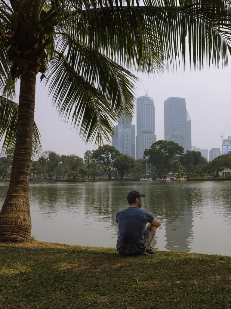 A man sitting alone on the grass beneath a palm tree, gazing across a lake toward a hazy Bangkok skyline with modern skyscrapers in Lumpini Park.