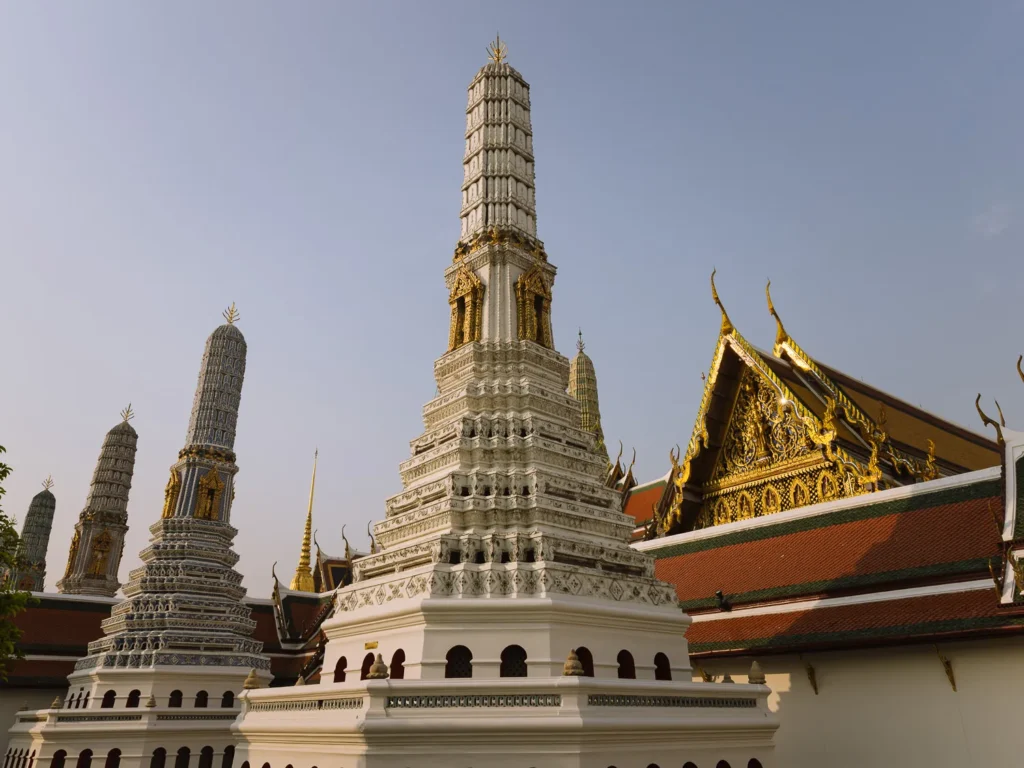 Tiered white chedi and golden roofs at Bangkok Grand Palace, Wat Phra Kaew temple against blue sky.