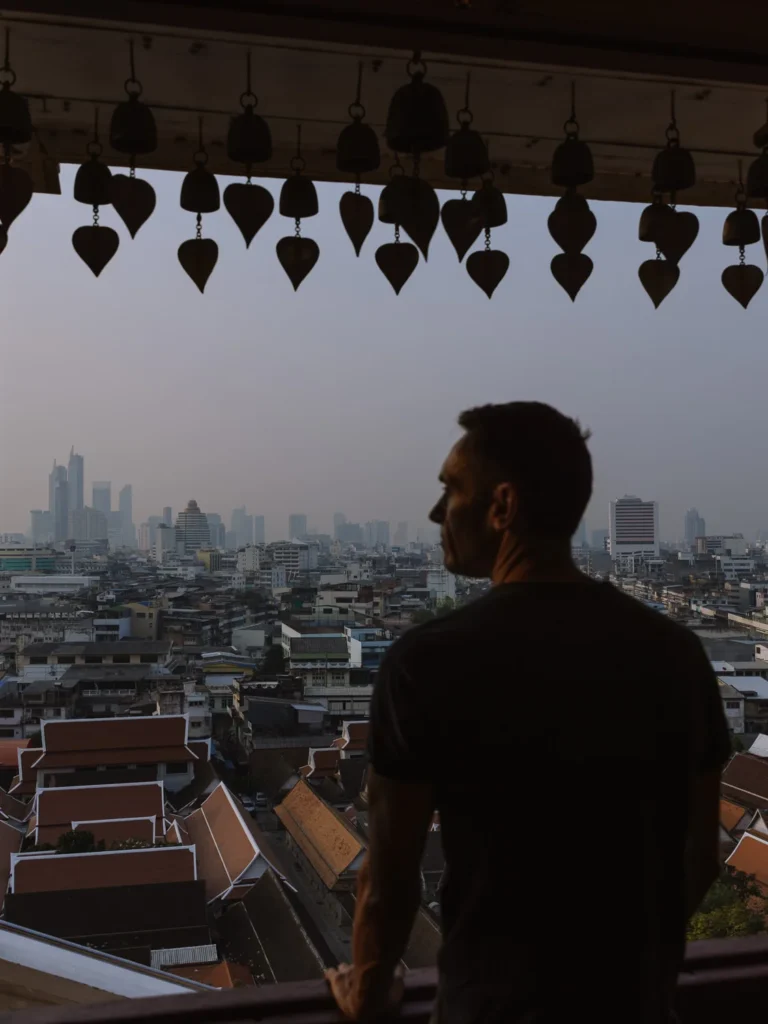 Silhouette of man gazes at hazy Bangkok skyline from Golden Mount balcony framed by leaf and bell ornament