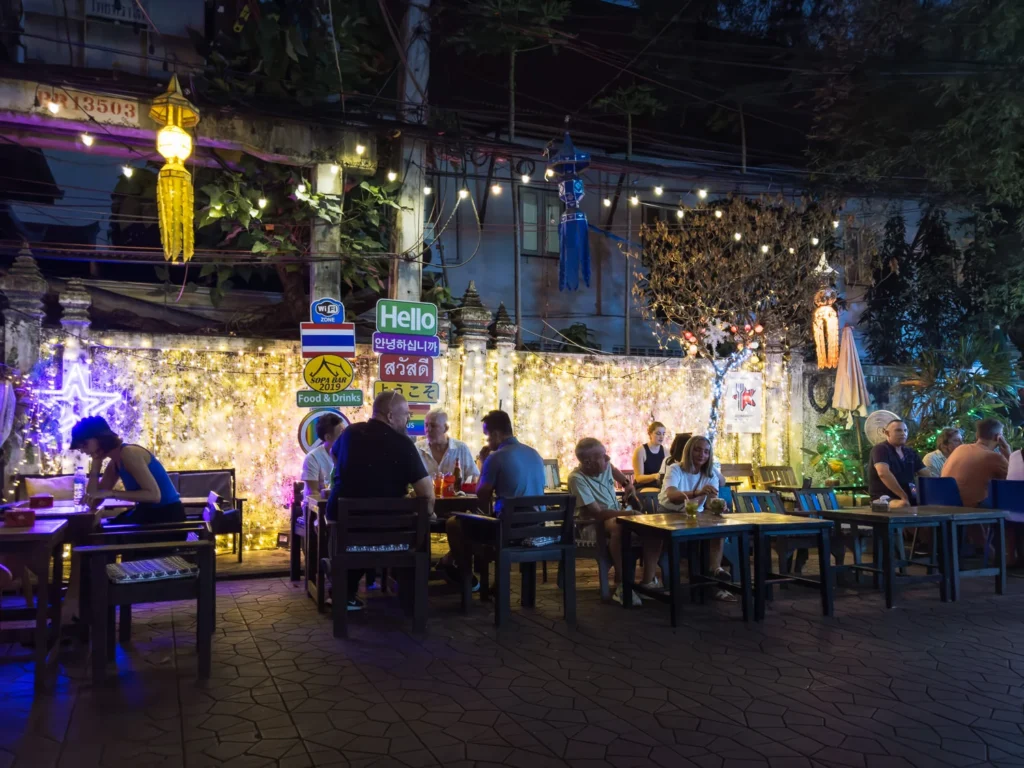Tourists dine outdoors at lively Khao San Road bar in Bangkok, lit by lanterns and fairy lights at night
