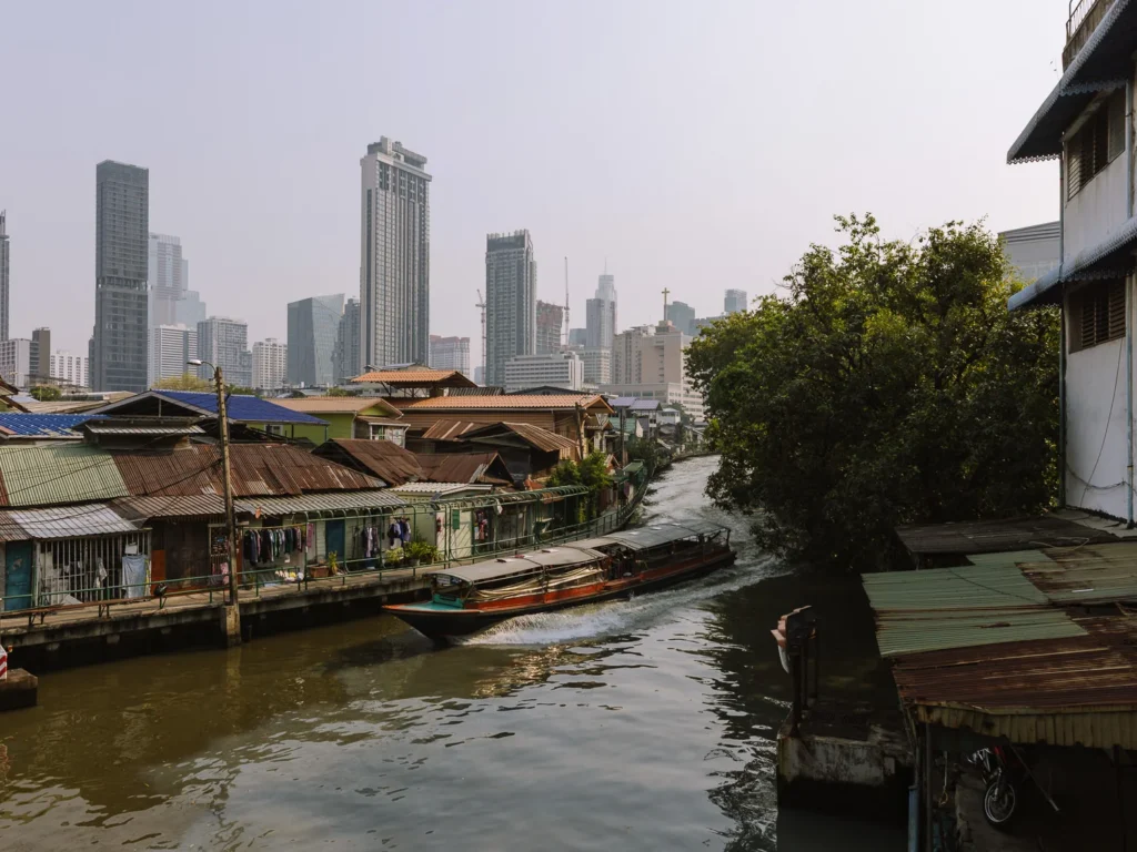 Longtail boat cruises Bangkok klong past riverside stilt houses toward modern skyscrapers.