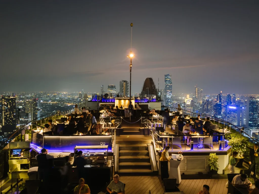 Rooftop bar overlooking Bangkok's glittering skyline at night, with patrons at lounges and blue-lit skyline.