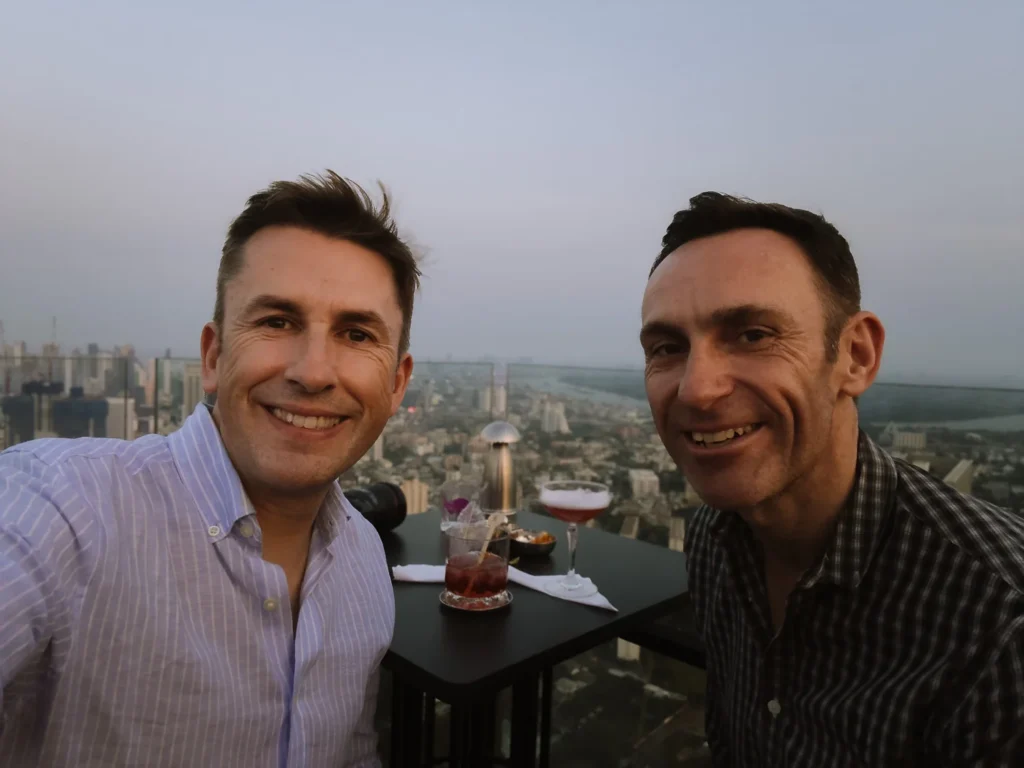 Two smiling men in button-up shirts toast cocktails at sunset on a Bangkok rooftop bar with skyline view.